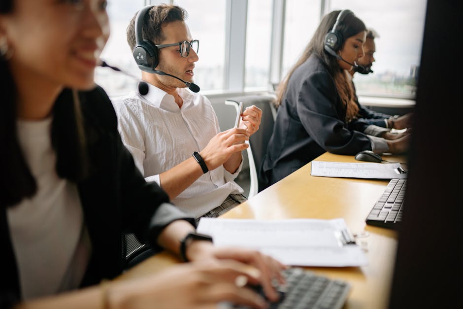 A group of customer service representatives in an office setting, wearing headsets and working at desks with keyboards, papers, and computers. Natural light from large windows illuminates the space, c