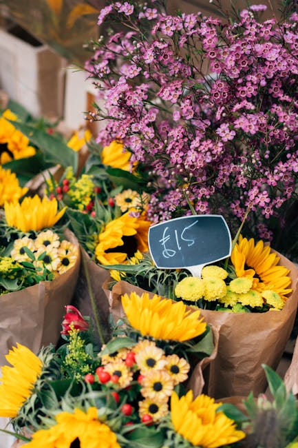 A colorful display of fresh flower bouquets at a flower market, featuring bright yellow sunflowers, small yellow daisies, and red accents wrapped in brown paper. In the background, there is a large cl