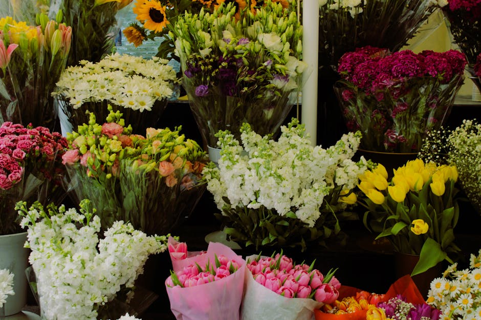 A diverse array of fresh flowers arranged in bouquets at a flower shop, featuring white daisies, pink tulips, purple lisianthus, yellow tulips, and clusters of pink roses. The flowers are wrapped in c
