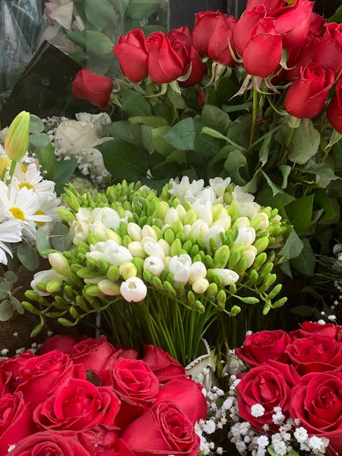 A vibrant floral arrangement featuring clusters of red roses with lush green foliage, complemented by white and yellow daisy-like flowers and a dense bundle of small white buds on long green stems. Th