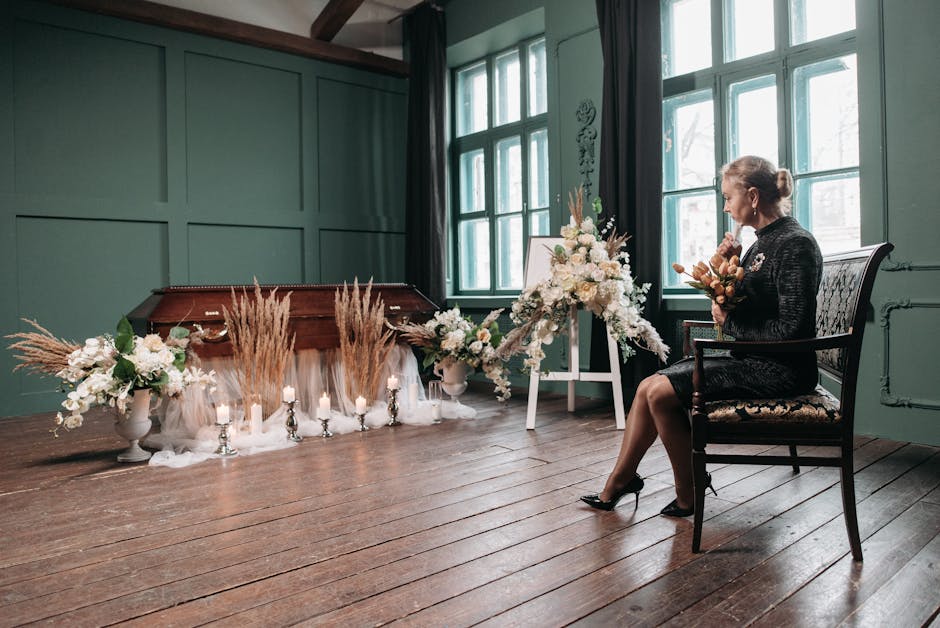 A woman dressed in black, sitting on an ornate wooden chair with upholstered seat, holds a small floral arrangement in her hands. In front of her, a large funeral wreath composed of white roses, lilie