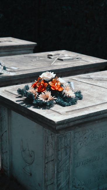 A floral wreath resting on a memorial stone, featuring a combination of fresh orange and white flowers, including daisies and roses, interwoven with dark green pine-like foliage. The arrangement is ci