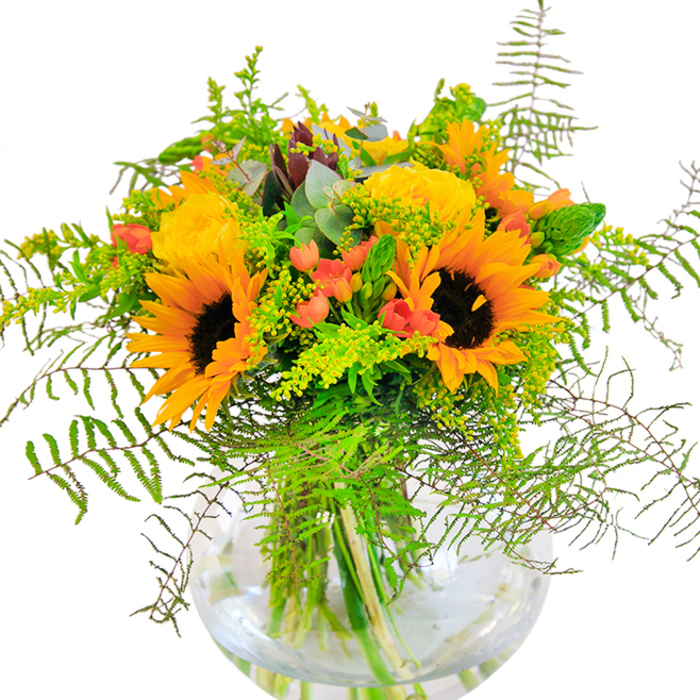 Pure sunlit joy spills from this dazzling floral arrangement, conceived by an artisan florist in Pinner and perfect for brightening special occasions or everyday moments. Golden yellow roses and radiant sunflowers capture the first impression, their petals unfurling in lush, overlapping spirals, almost seeming to pulse with summery warmth reminiscent of mornings in Pinner Memorial Park. Around them, cascades of deep green foliage and soft, stone-blue eucalyptus leaves arch gracefully outward, the feathery textures recalling quiet walks along the leafy avenues near Love Lane. Glossy red Hypericum berries dot the bouquet with vivid, jewel-like contrasts, their smooth skins gleaming against the matte, velvety sunflower faces. Held within a gleaming, rounded glass vase, the bouquet's fresh-cut stems are visible-a testament to the daily freshness and care local florists in Pinner provide their clientele. Up close, one can sense the interplay of textures: petals like silk, foliage crisp and lively, the light fragrance of roses and greenery conjuring open air and sunny paths. Brilliant in the soft daylight, this bouquet becomes an ode to happiness, perfect for birthdays, anniversaries, or any event where celebration and heartfelt joy are essential to the moment.